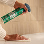 A person uses one hand to spray Grove Co. Multi-Purpose Cleaner – Winter Collection onto a white surface and wipes it clean with a cloth, set against light-tiled walls in the background.