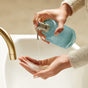 A person pumps foam from the Grove Co. Hand Soap Dispenser - Winter Collection into their hand over a white sink with a gold faucet.