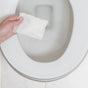 A hand holds a white sanitary pad over an open, white toilet with a tiled background. Nearby are Grove Co. Toilet Bowl Cleaner Pods, designed to tackle hard water stains and leave behind a fresh wild mint scent.
