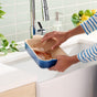 A person cleans a blue baking dish with Grove Co. Gentle Walnut Scouring Pads under a kitchen sink faucet, while soap and citrus fruits sit in the background.