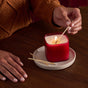 A person in a brown sweater lights a Grove Co. Candles 9.5oz - Fall Collection candle, made with upcycled ingredients, on a white plate with matches on a wooden table.