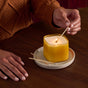 A person in a brown sweater lights the eco-friendly Grove Co. Candle - Fall Collection (white pumpkin & ginger scent), in a yellow square jar on a white plate, with matches scattered on a wooden table for cozy autumn vibes.