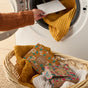 Wearing an orange sweater, a person adds a Grove Co. Laundry Sheets - Fall Collection sheet into a washer with a yellow textured blanket. In front, a basket holds more laundry and two boxes of the Grove Co. detergent sheets.