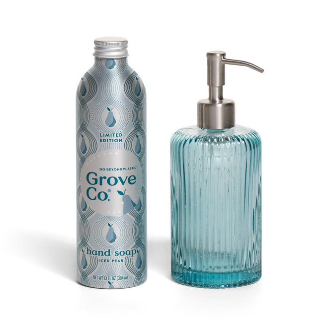 A Grove Co. Hand Soap Starter Set - Winter Collection canister stands next to a clear, ribbed blue glass soap dispenser with a silver pump.