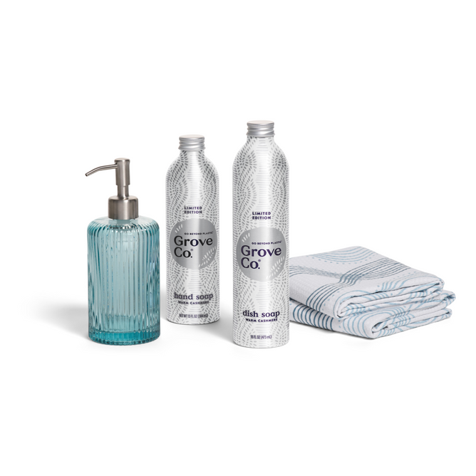 The Grove Co. Hygge Home Set - Winter Collection features a blue glass soap dispenser, two labeled bottles for hand and dish soap, and two blue-and-white patterned towels, all arranged on a white background.
