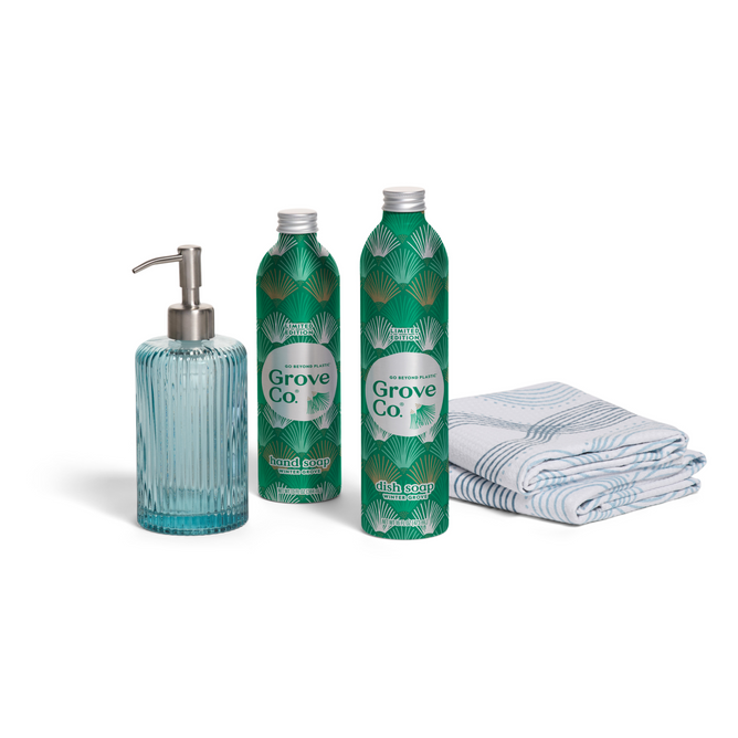 The Grove Co. Hygge Home Set - Winter Collection features a ribbed blue glass soap dispenser, two green dish soap refill bottles with leaf patterns, and a folded white and blue patterned dish towel on a white background.