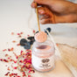 A hand holds a gold spoon with pink powder above an open Anima Mundi ROSE Face Mask jar. Surrounded by dried rose petals, this Detoxify + Hydrate clay mask offers a luxurious, botanical skincare experience on a white surface.