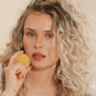 A woman with long, curly blonde hair and blue eyes holds the yellow, round Hydrating Conditioner Bar from Good Juju Body & Home near her face. She has light skin and natural makeup against a soft beige background.