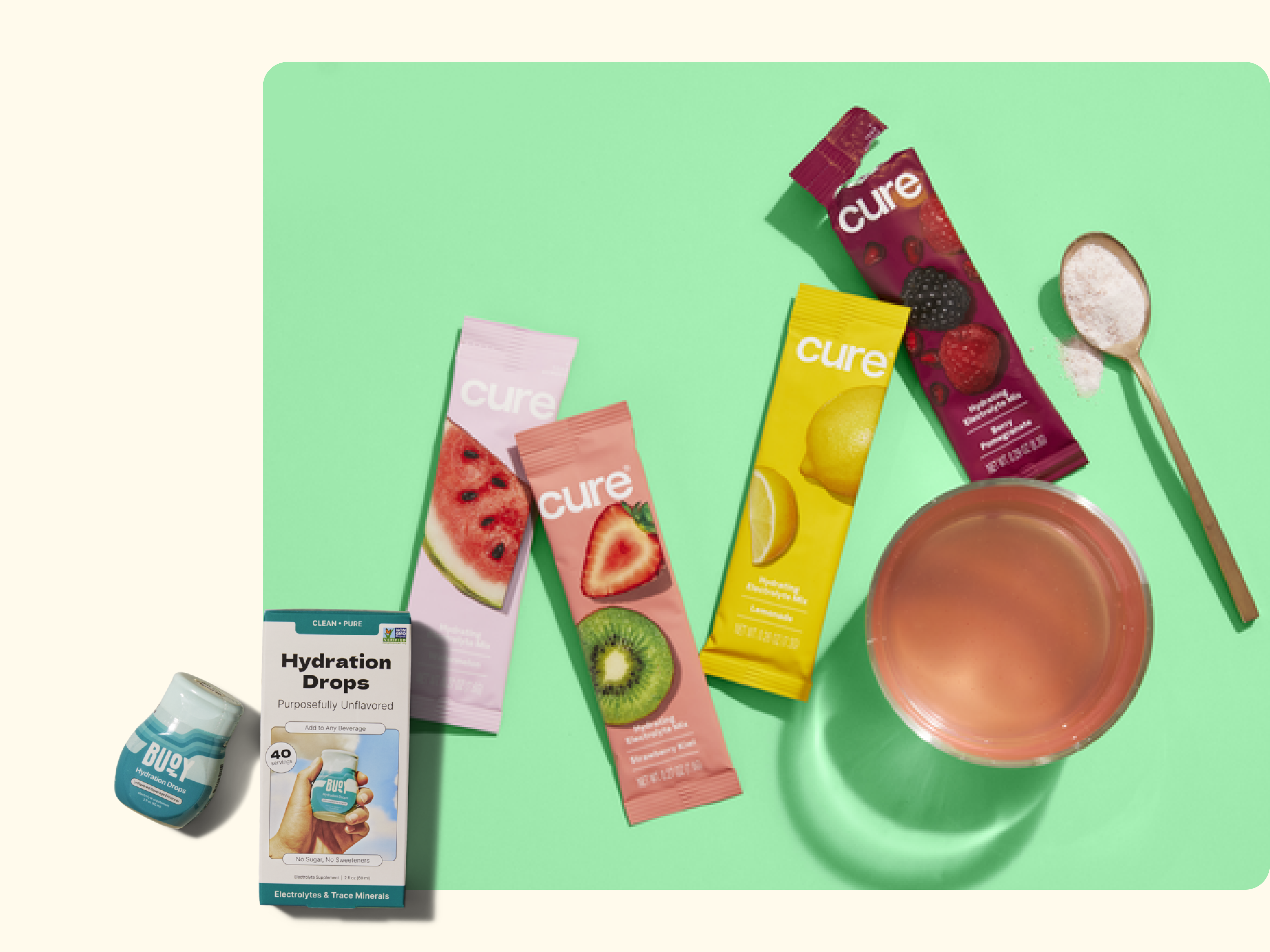 Assorted Cure hydration drink mix packets and a glass of pink beverage are displayed on a green background, with a wooden spoon containing powder and a bottle and box of hydration drops nearby.