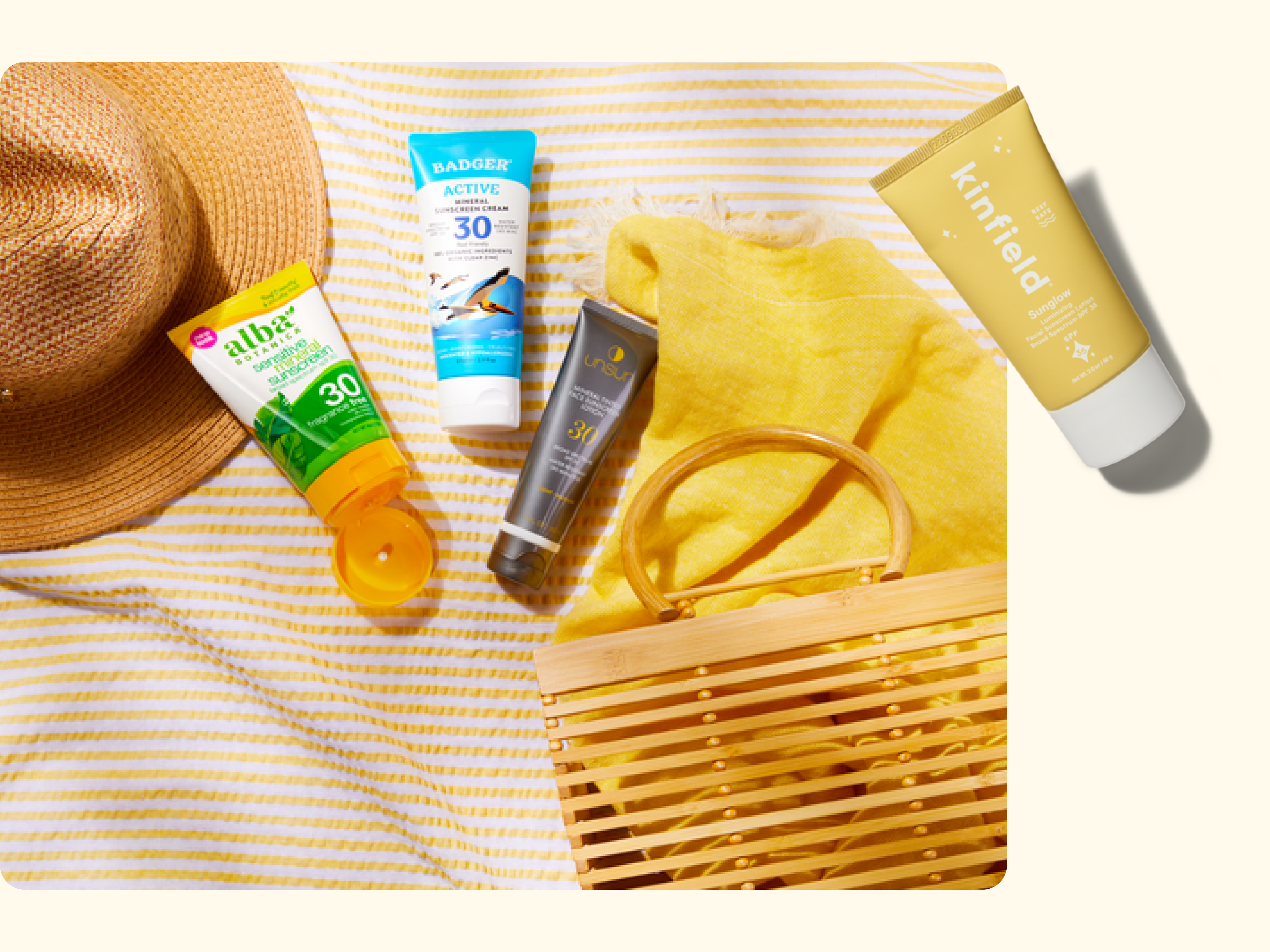 A straw sunhat, striped yellow towel, bamboo handbag, and four sunscreen bottles arranged on a yellow and white striped surface, suggesting a summer or beach day.