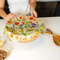 A person covers a wooden bowl with Bees Wrap Assorted Wraps-5 pack on a white kitchen counter, surrounded by small bowls of snacks and wooden utensils, showcasing the eco-friendly convenience of these reusable food wraps.