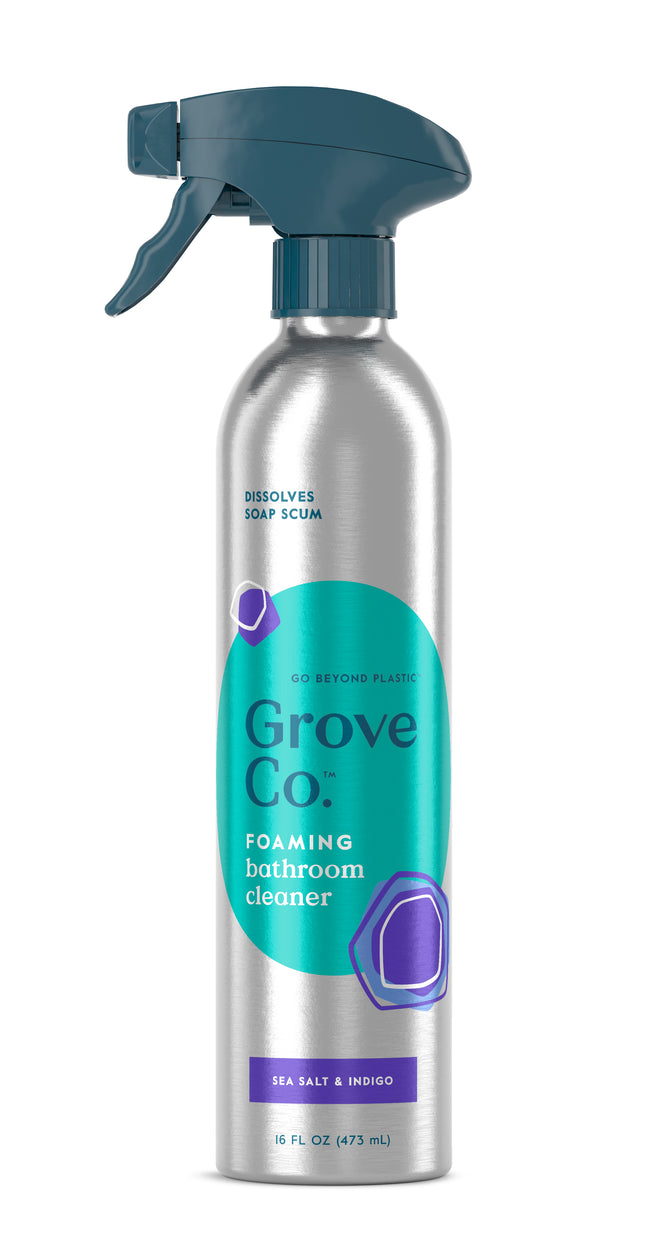 A recyclable silver aluminum spray bottle of Grove Co. Foaming Bathroom Cleaner, featuring a teal nozzle and label reading: Dissolves soap scum with a multi-purpose foaming formula enhanced by Sea Salt & Indigo. The bottle holds 16 fl oz (473 ml).