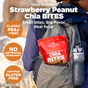 A hiker's backpack outdoors features a bright red Navitas Organics Superfood+ Strawberry Peanut Chia BITES bag—tasty, certified gluten-free peanut butter bites with PB&J flavor and no artificial ingredients.
