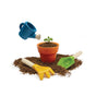A small plant grows in an orange pot with soil, while a blue watering can, yellow rake, and green shovel from the PlanToys Gardening Set lie nearby—ideal for outdoor play.