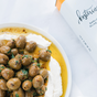 A plate of green olives in seasoned extra virgin olive oil rests on whipped cheese, beside a Kosterina Garlic Olive Oil container.