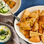A white platter of toasted pita chips brushed with Kosterina Garlic Olive Oil and herbs sits next to cucumber yogurt dip with dill; a small plate with dip and chips rests on a blue textured placemat and gray napkin.
