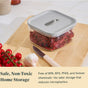 A Caraway Glass Airtight Container Set with a gray lid sits on a wooden counter, filled with dried chili peppers. Cherry tomatoes, garlic, and a knife are nearby. The text emphasizes the set's chemical-free design for safe kitchen storage.