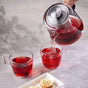 A hand pours vibrant red tea from a SAKI Electric Samovar into a cup beside another already filled cup. In the foreground, three round, nut-covered cookies rest on a white rectangular plate, creating an inviting scene perfect for keeping warm on chilly days.