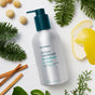 A bottle of Ursa Major Go Easy Conditioner, known for revitalizing and volumizing hair, is displayed on a white background with pine sprigs, lemon, cinnamon sticks, macadamia nuts, and green leaves around it.