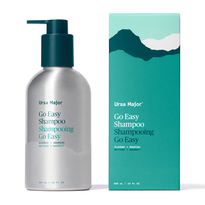 A silver pump bottle and matching teal box of Ursa Major Go Easy Shampoo, a daily shampoo for all hair types, are shown side by side. The box displays a mountain design with product information in teal, white, and yellow.