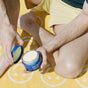 A person in yellow shorts sits on a yellow towel, holding Ursa Major’s Golden Hour Body Butter—open blue jar in hand—ready to restore dry skin with deep hydration.