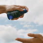 A hand pours Ursa Major’s Golden Hour Dry Oil from a blue bottle onto another hand, with a blue sky backdrop—perfect for locking in moisture and replenishing skin and hair.