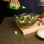 Someone pours Graza Drizzle Glass Extra Virgin Olive Oil, made from early harvest olives, over a bowl of mixed greens and radishes on the kitchen counter, with extra radishes, a cutting board, and a plate nearby.
