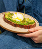 A hand holds a ceramic plate with avocado toast and poached egg, dressed in blue denim, drizzling Graza Drizzle Glass Extra Virgin Olive Oil—made from early harvest olives and rich in antioxidants—over the dish.