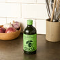 A green bottle of Graza Drizzle Glass Extra Virgin Olive Oil is on a kitchen countertop beside a bowl of garlic and shallots, with a utensil holder in the background.