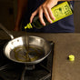 Someone pours Graza Sizzle Glass Extra Virgin Olive Oil from a green bottle into a stainless steel frying pan on the stove, filling the kitchen with olive aroma; its yellow cap lies on the counter nearby.