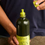 A person in a navy blue shirt holds a bottle of Graza Sizzle Glass Extra Virgin Olive Oil, unscrewing the green cap to reveal the pour spout. This olive oil offers a mellow flavor that complements any dish.