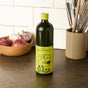 A bottle of Graza Sizzle Glass Extra Virgin Olive Oil rests on a kitchen counter, ideal for everyday cooking. Nearby are a bowl with garlic and onions and a container filled with kitchen utensils.