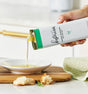 A hand pours Kosterina Green Goddess Olive Oil from a rectangular tin into a white dish on a wooden board, with bread pieces nearby and a green textured cloth in the background.