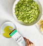 A hand pours Kosterina’s Green Goddess Olive Oil from a white and teal bottle into a small dish, with chopped green vegetables and tortilla chips arranged nearby on a wooden board.