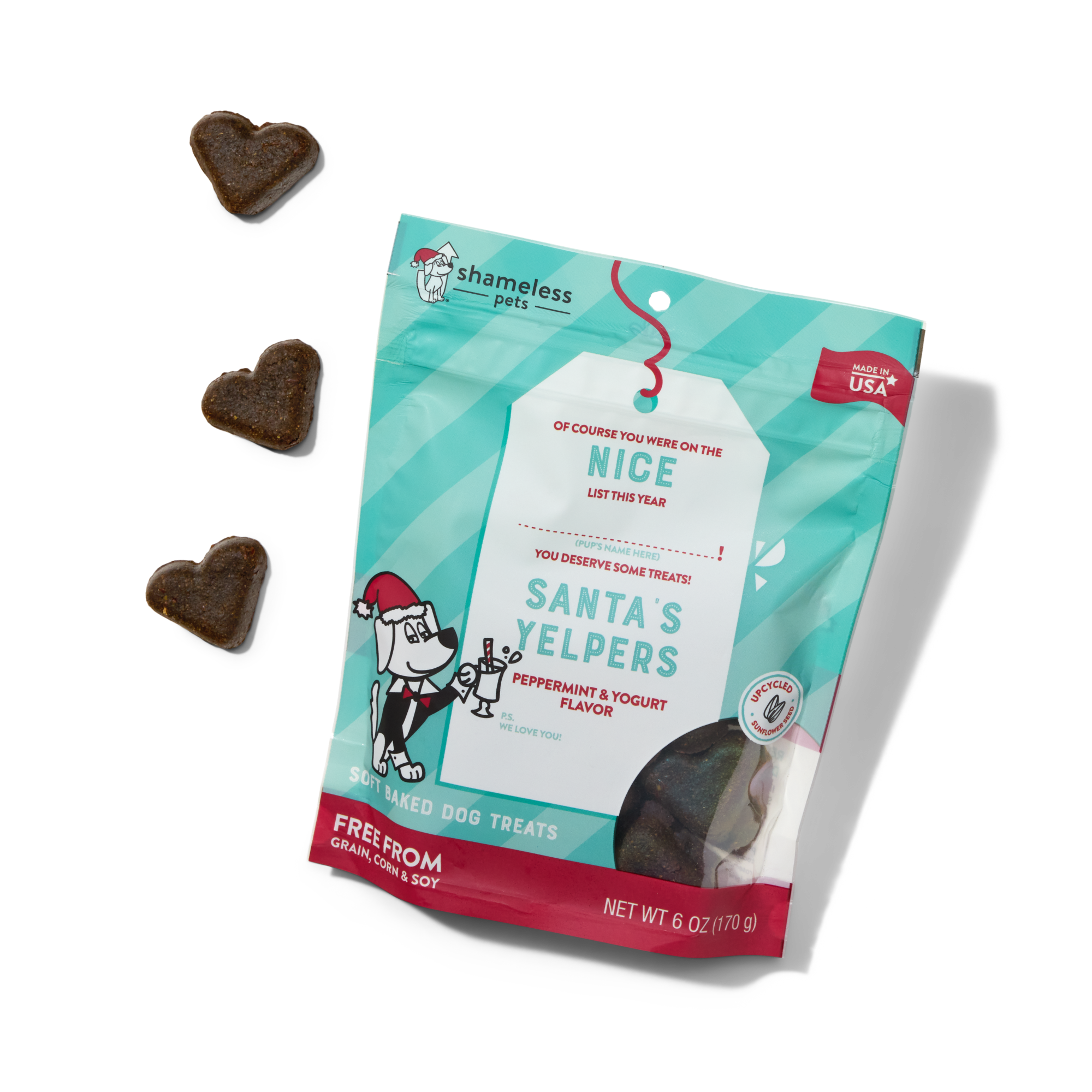 A teal and red bag of Shameless Pets Santa’s Yelpers dog treats (peppermint and yogurt flavor) lies on a white background, with three heart-shaped treats beside it. The packaging features a festive design with a dog illustration.