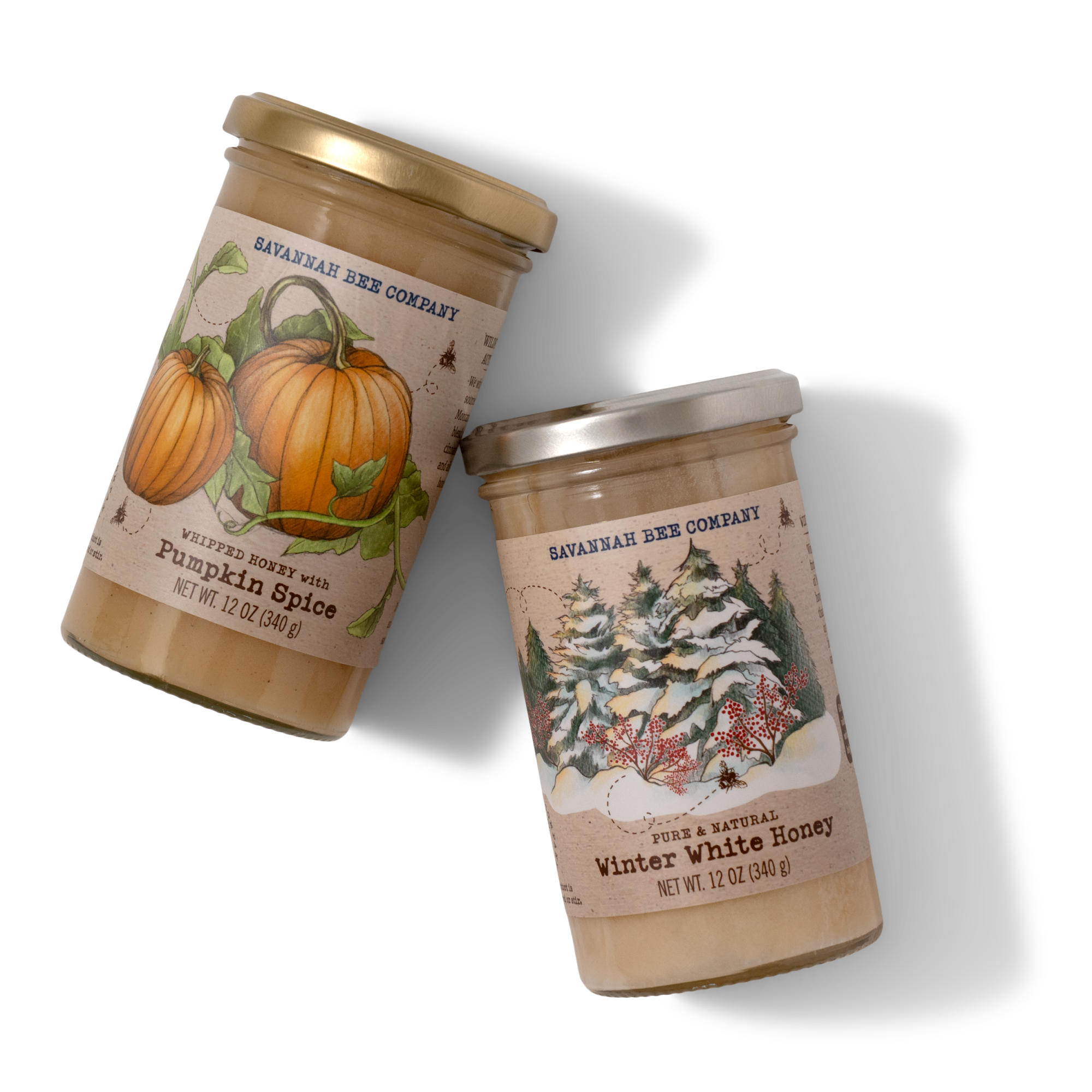 Two jars of Savannah Bee Company whipped honey, one labeled Pumpkin Spice with a pumpkin illustration, and the other labeled Winter White Honey with a snowy evergreen scene, both with gold lids.