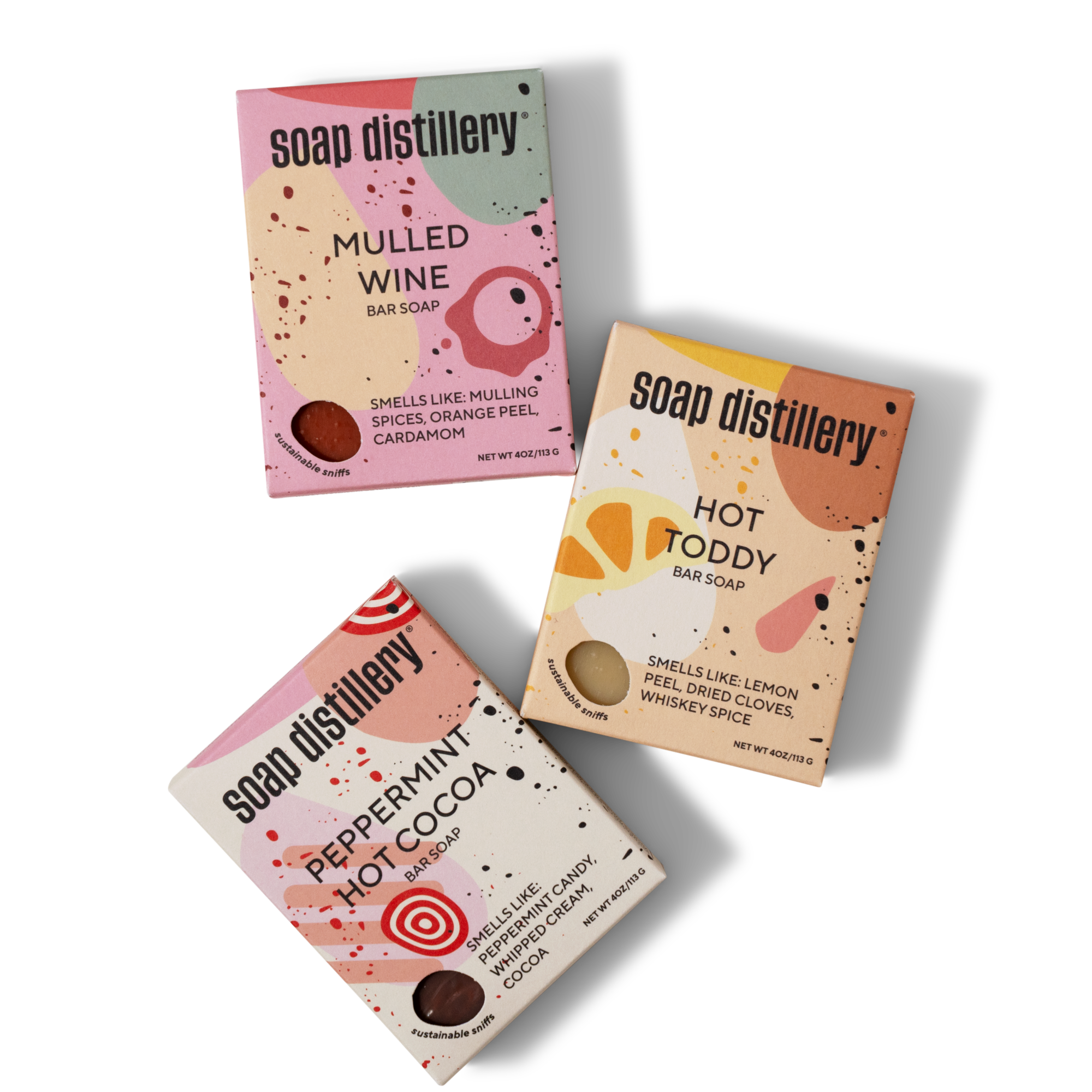 Three rectangular soap boxes from Soap Distillery in Mulled Wine, Hot Toddy, and Peppermint Hot Cocoa scents, each with colorful abstract designs and small circular windows showing the soap inside.