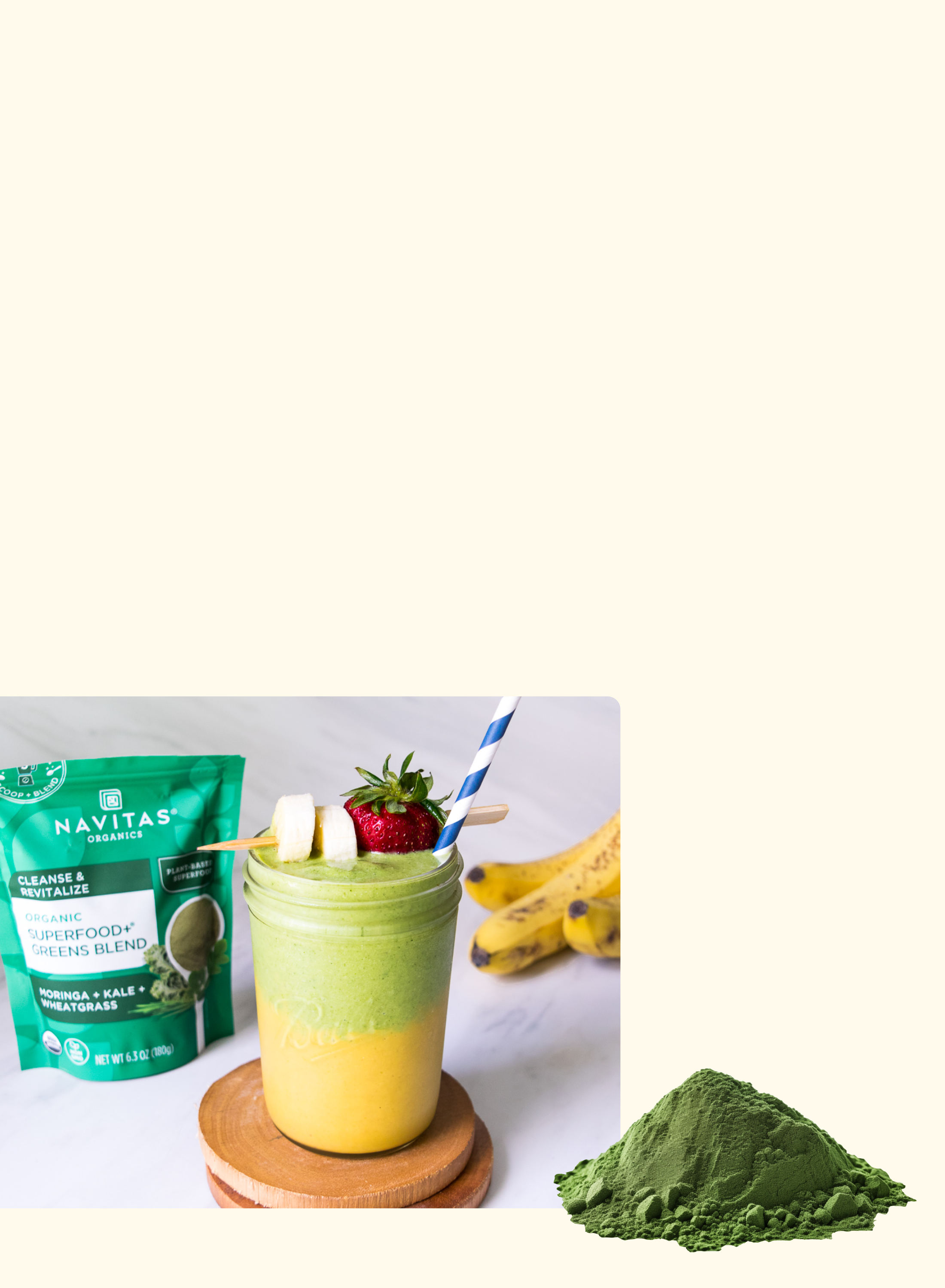 A layered green and yellow smoothie in a jar, topped with banana slices and strawberries, sits beside a striped straw, a bunch of bananas, a packet of Navitas Superfood Greens Blend, and a heap of green powder.