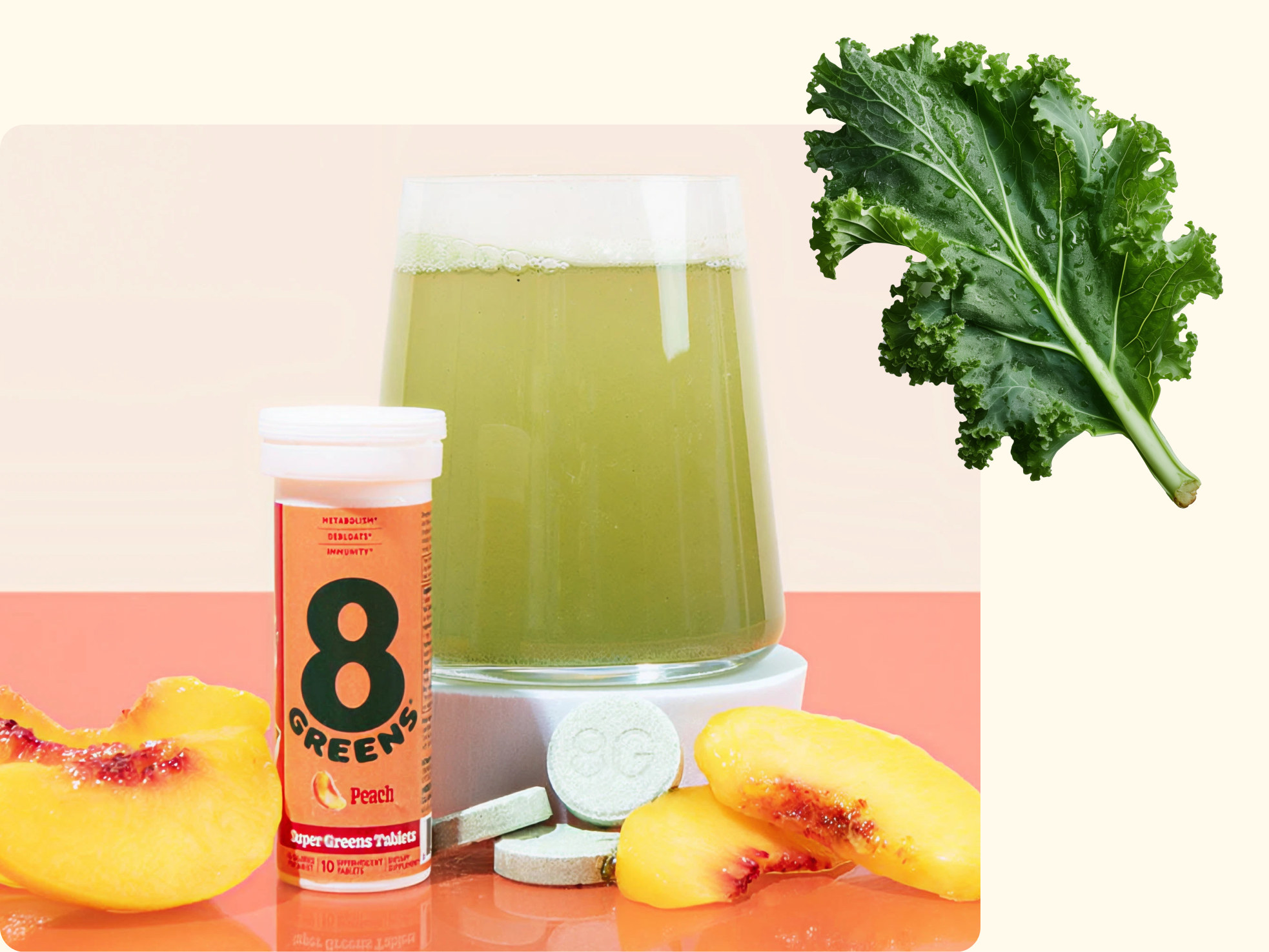 A glass of green drink sits next to a tube of 8 Greens peach tablets, two green tablets, peach slices, and a kale leaf on a light background.