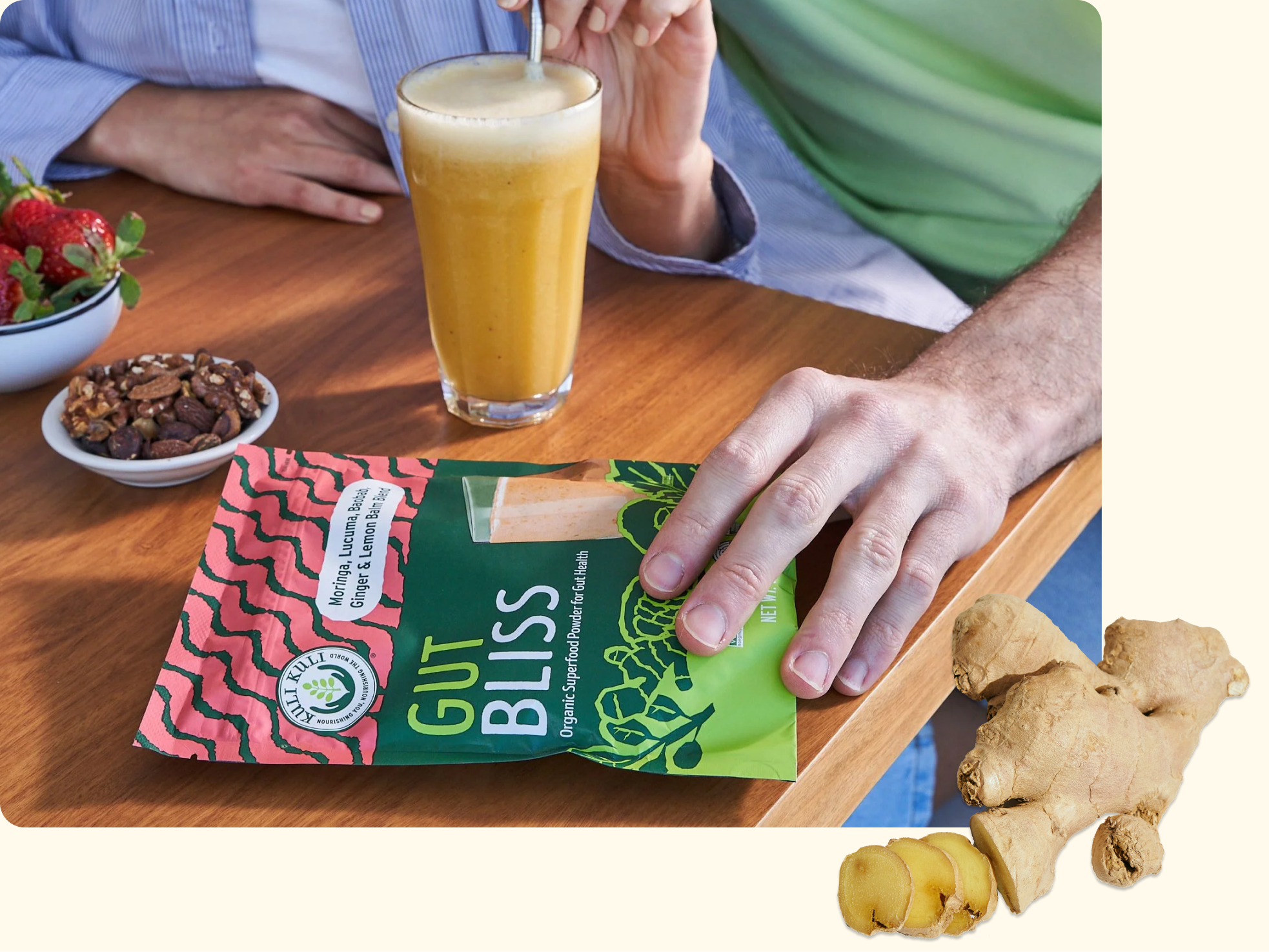 A person holds a Gut Bliss supplement packet next to a glass of smoothie on a wooden table, with a bowl of nuts and strawberries nearby and fresh ginger root in the foreground.