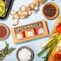 A Spicewalla Spices & Seasonings - Grill & Roast Collection set is surrounded by fresh garlic, rosemary, green onions, peppercorns, tomatoes, mustard seeds, and a bowl of salt on a blue-tiled surface.