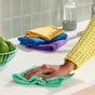 A person in a yellow sweater wipes a white countertop with a green Grove Co. Assorted Cleaning Cloth from the 4 pk set. In the background are folded blue, purple, and yellow cloths, a bowl of limes, and a cleaning spray bottle.