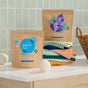 Two Grove Co. Laundry Detergent Packs, 45 count, are placed on a laundry room counter near stacked colorful towels, wool dryer balls, a wicker basket, and green tiles in the background.