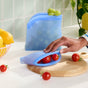 A hand places cherry tomatoes into a Grove Co. Reusable Silicone Bag on a wooden cutting board, with another sustainable silicone bag of green grapes and a bowl of lemons in the background.