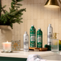 A bathroom counter featuring Grove Co. Hand Soap Refill - Winter Collection bottles, a lit candle, a box of plant-based hand soap with natural fragrance, a glass soap dispenser, a towel, a green apple, and a pine branch arranged in a vase—all set against tiled walls illuminated by warm lighting.