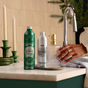A hand presses the pump on a clear soap dispenser at the sink, alongside Grove Co. dish soap and Grove Co. Hand Soap Refill from the Winter Collection. The scene features green and white decor, candles, and other seasonal accents in the background.