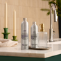 Two Grove Co. Hand Soap Refill bottles from the Winter Collection and a ribbed glass soap dispenser sit on a marble countertop near a sink, with green candle holders and a folded towel in the background. The arrangement suggests plant-based hand soap with natural origin fragrance, offering an elevated kitchen experience.
