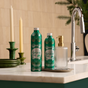 Two green Grove Co. Hand Soap Refill bottles from the Winter Collection sit on a marble counter beside a ribbed glass soap dispenser with a gold pump. Green candlesticks and a tall faucet are in the background, adding to the cozy charm of this collection inspired by natural origin fragrances.
