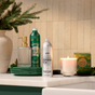 Green and silver bottles of Grove Co. Hand Soap Refill – Winter Collection, featuring a plant-based formula with a natural origin fragrance, sit alongside a glass soap dispenser, a lit candle, and a green box on a white bathroom countertop with stacked green plates and a vase of fir branches in the background.