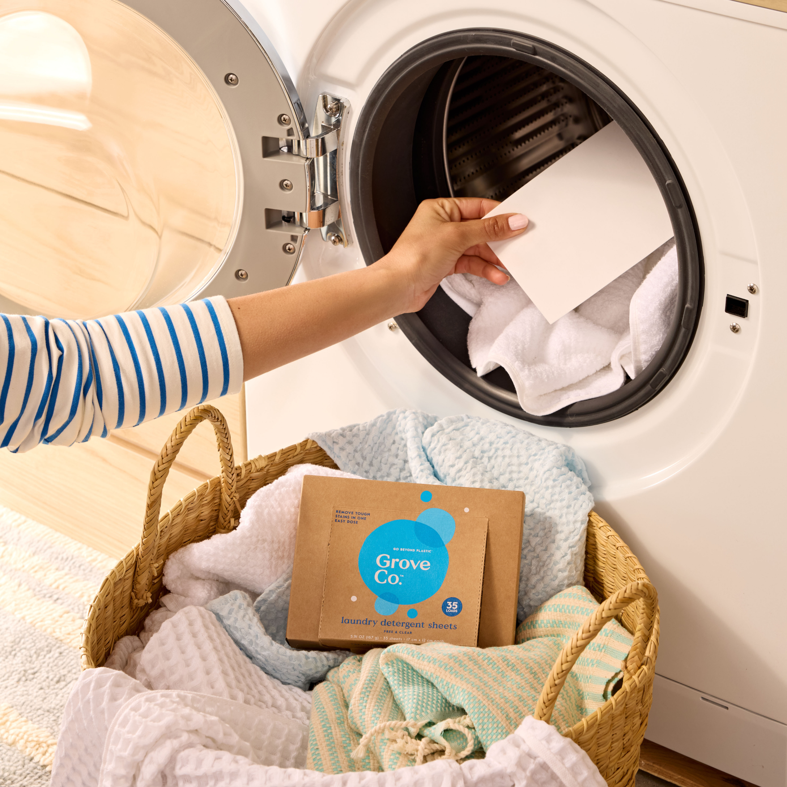 A person loads a Grove Co. Laundry Detergent Sheet into a front-loading washer, with a basket of towels and the eco-friendly detergent box highlighting their choice to reduce plastic waste.